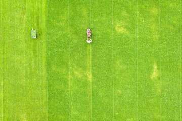 Aerial view of small figure of man worker trimming green grass with mowing mashine on football stadium field in summer.