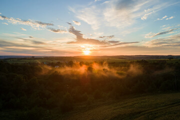 Aerial view of light fog covering dark forest trees at warm sunset.