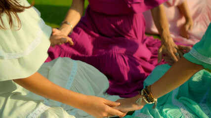 Obraz premium European girls friends in dresses sitting in a circle holding each other's hands in nature with green grass on the background