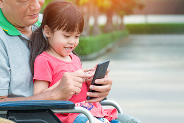 Happy Asian disabled man and his granddaughter on wheelchair using smartphone togetherness while relaxing in public park area