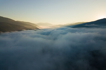 Aerial view of vibrant sunset over white dense foggy clouds with distant dark silhouettes of mountains on horizon.