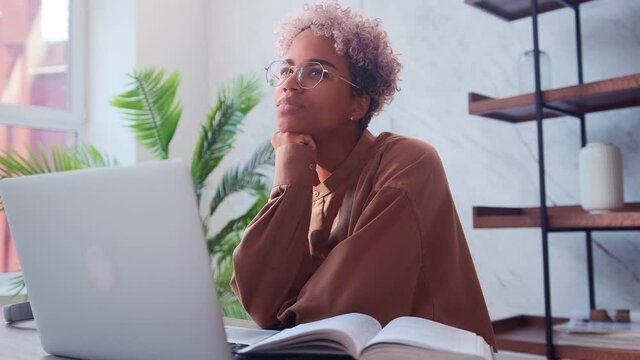 Beautiful dark skinned woman dreams of something while sitting on laptop in office. Young woman ponders new ideas or dreams of positive things while being distracted from work. Daydreaming concept.