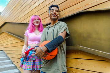 Fototapeta na wymiar alternative diverse couple hanging out together chilling and smiling standing against background wall. happy interracial friends holding skateboard and basketball relaxing outdoors. lifestyle concept