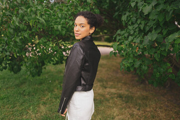 Fashion portrait of attractive confident young natural beauty African American woman with afro hair in black leather jacket posing in nature park in green foliage.