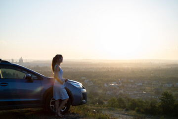 Fototapeta premium Happy young woman driver in blue dress leaning on her car enjoying warm summer day. Travelling and vacation concept.