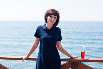 A smiling brunette with a glass of juice is standing on pier by the sea.