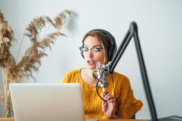 Charismatic woman radio host recording podcast at home studio. Female podcaster recording broadcasting into microphone using laptop
