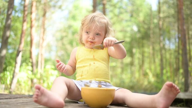 Little funny cute blonde girl child toddler with dirty clothes and face eating baby food fruit or vegetable puree with spoon from yellow plate outside at summer. Healthy happy childhood concept