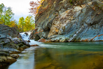 Mountain river with small waterfall with clear turquoise water falling down between wet boulders with thick white foam on autumn bright day.