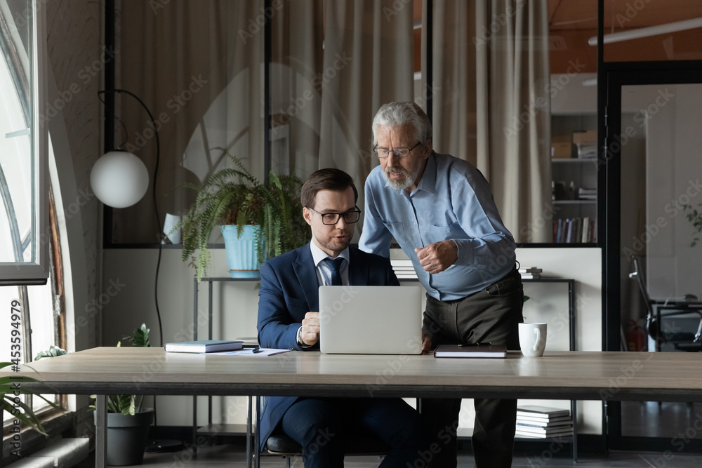 Poster senior businessman wearing glasses mentor instructing training new employee, using laptop, helping w