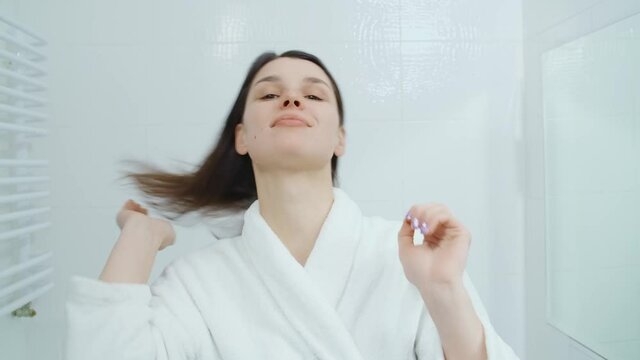 Front view of woman after shower in white bathrobe admiring appearance in modern mirror and braiding thick hair in ponytail. Concept of woman admiring appearance in bathroom.