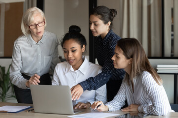 Confident diverse businesswomen colleagues discussing project, looking at laptop screen, employees workers with mature team leader executive brainstorming, sharing ideas, discussing strategy