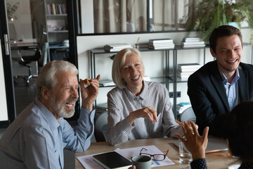 Excited mature businesspeople with colleagues eating pizza during break in office together, happy senior coworkers laughing at funny joke, talking chatting having fun, sharing corporate lunch