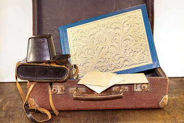 An old vintage brown suitcase with a retro camera in a leather case, an album with photos on a wooden table - Powered by Adobe