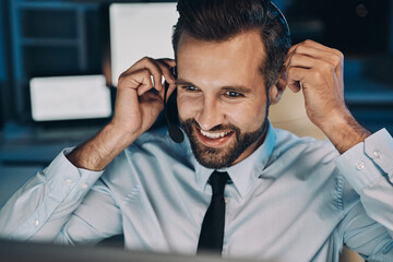 Happy young man in headset looking at the computer and smiling while staying late in the office