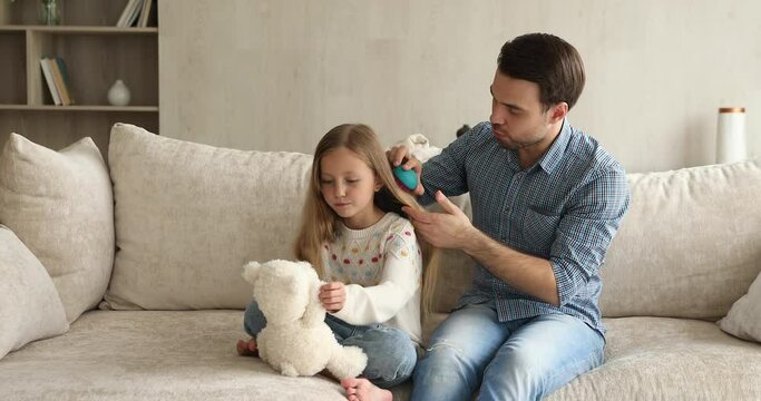 Cute little 9s girl sit on sofa play fluffy bear while young single daddy combing her long hair with modern hairbrush, making hairdo. Family routine, take care of daughter, happy fatherhood concept