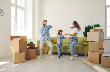 Fototapeta na wymiar Happy young family having fun in their new home on moving day. Cheerful excited mum, dad and child playing and dancing in room with sofa and unpacked boxes. Real estate, mortgage, buying house concept
