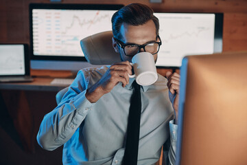 Confident young man drinking coffee and working on computer while staying late in the office