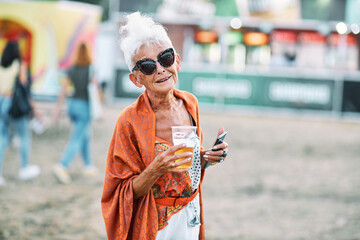 An old woman with gray hair is standing outdoor, drinking beer and having a great time at the festival. - obrazy, fototapety, plakaty