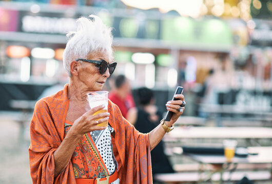 An Old Woman With Short Gray Hair Is Standing Outdoor At A Music Festival, Drinking Beer And Using Her Phone. Elders Young In Heart.