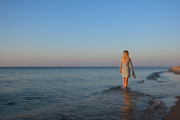 A young blonde girl in the morning goes on the beach barefoot and carries shoes in her hand. Back view