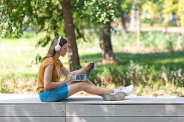 Digital nomad. Caucasian woman in wireless headphones sits on a parapet and working on a laptop in the public park outdoor in summer, side view, selected focus.