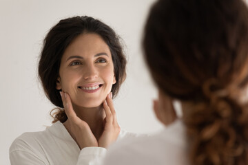 Head shot attractive happy young hispanic latina woman in robe looking in mirror, touching face, enjoying morning evening anti aging skincare routine, feeling satisfied with perfect skin condition.