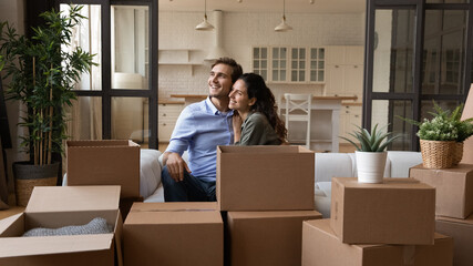 Dreamy young happy bonding family couple cuddling sitting on sofa near big carton boxes, visualizing future, planning decoration in renovated stylish living room, resting together in own home.