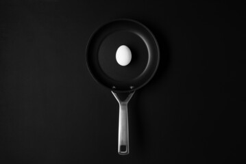 Empty black frying pan on black wooden table background with white raw egg. Creative food art cooking