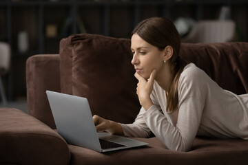 Fototapeta premium Close up side view thoughtful focused young woman looking at laptop screen, lying on couch at home, pensive female businesswoman freelancer or student pondering project strategy, reading news