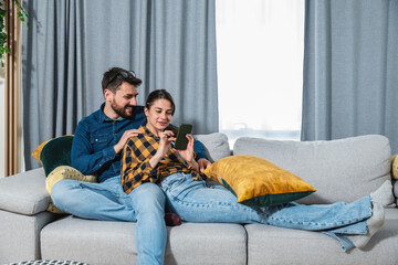 Young happy couple man and woman relaxing on the sofa in their home surfing the internet on the smartphone looking for funny videos and memes to have quality free time on weekend  - obrazy, fototapety, plakaty