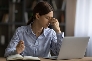 Exhausted young woman taking off glasses, feeling eye strain, sitting at work desk with laptop,...