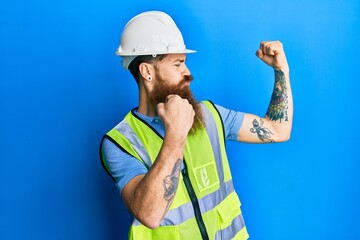 Redhead man with long beard wearing safety helmet and reflective jacket showing arms muscles...