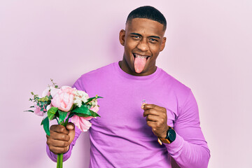 Young black man holding bouquet of flowers and wedding ring sticking tongue out happy with funny expression.