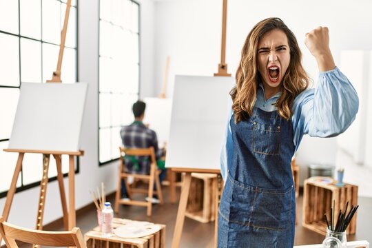 Young artist woman at art studio angry and mad raising fist frustrated and furious while shouting with anger. rage and aggressive concept.