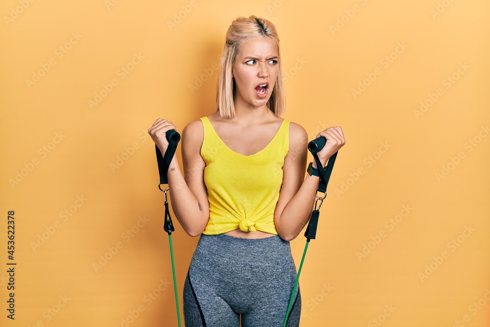 Poster beautiful blonde woman training arm resistance with elastic arm bands angry and mad screaming frustr