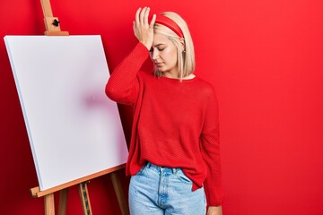 Beautiful blonde woman standing by painter easel stand surprised with hand on head for mistake,...