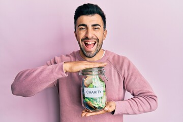 Young hispanic man holding charity jar with money smiling and laughing hard out loud because funny crazy joke.