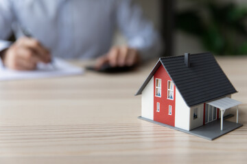 Close up of small toy miniature model of house on table. Homeowner, property buyer, realtor, mortgage broker calculating in background, counting loan, signing document, agreement. Real estate concept