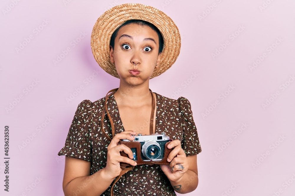 Poster beautiful hispanic woman with short hair wearing summer hat holding vintage camera puffing cheeks wi