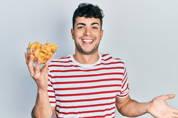 Young hispanic man holding bowl with uncooked pasta celebrating achievement with happy smile and winner expression with raised hand