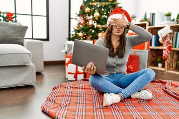 Young latin woman using laptop sitting by christmas tree covering eyes with hand, looking serious and sad. sightless, hiding and rejection concept