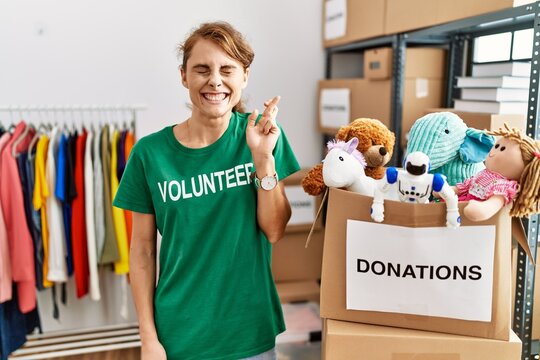 Beautiful caucasian woman wearing volunteer t shirt at donations stand gesturing finger crossed smiling with hope and eyes closed. luck and superstitious concept.