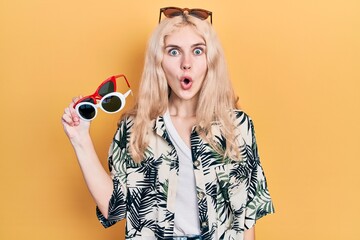 Beautiful caucasian woman with blond hair holding many sunglasses scared and amazed with open mouth for surprise, disbelief face