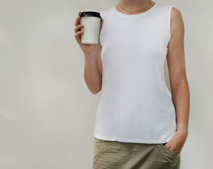 Close up of young girl in white tank top holding blank take away coffee cup in her hands over light gray background. Mockup of coffee to go cup and t-shirt print. Copy space for your branding design.