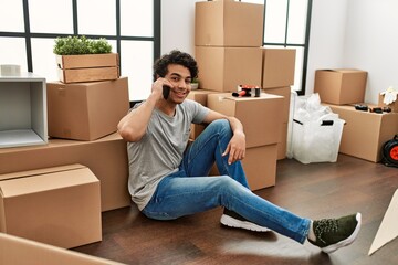 Young hispanic man talking on the smartphone sitting on the floor at new home.