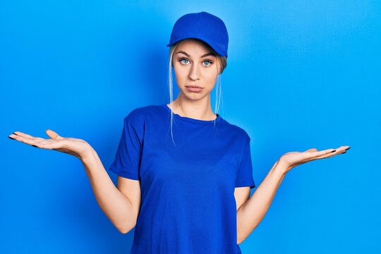 Young caucasian woman wearing courier uniform wearing cap clueless and confused expression with arms and hands raised. doubt concept.