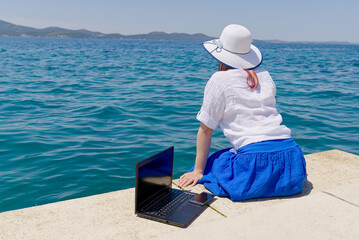 Work from anywhere. Side view of young woman, female freelancer in straw hat working on laptop while sitting on the beach. young woman in sitting on stone at sea and remotely working on laptop