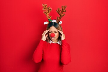 Beautiful hispanic woman wearing deer christmas hat and red nose with hand on head for pain in head because stress. suffering migraine.