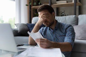 Frustrated desperate millennial man checking bills for payments, holding receipts, getting upset...